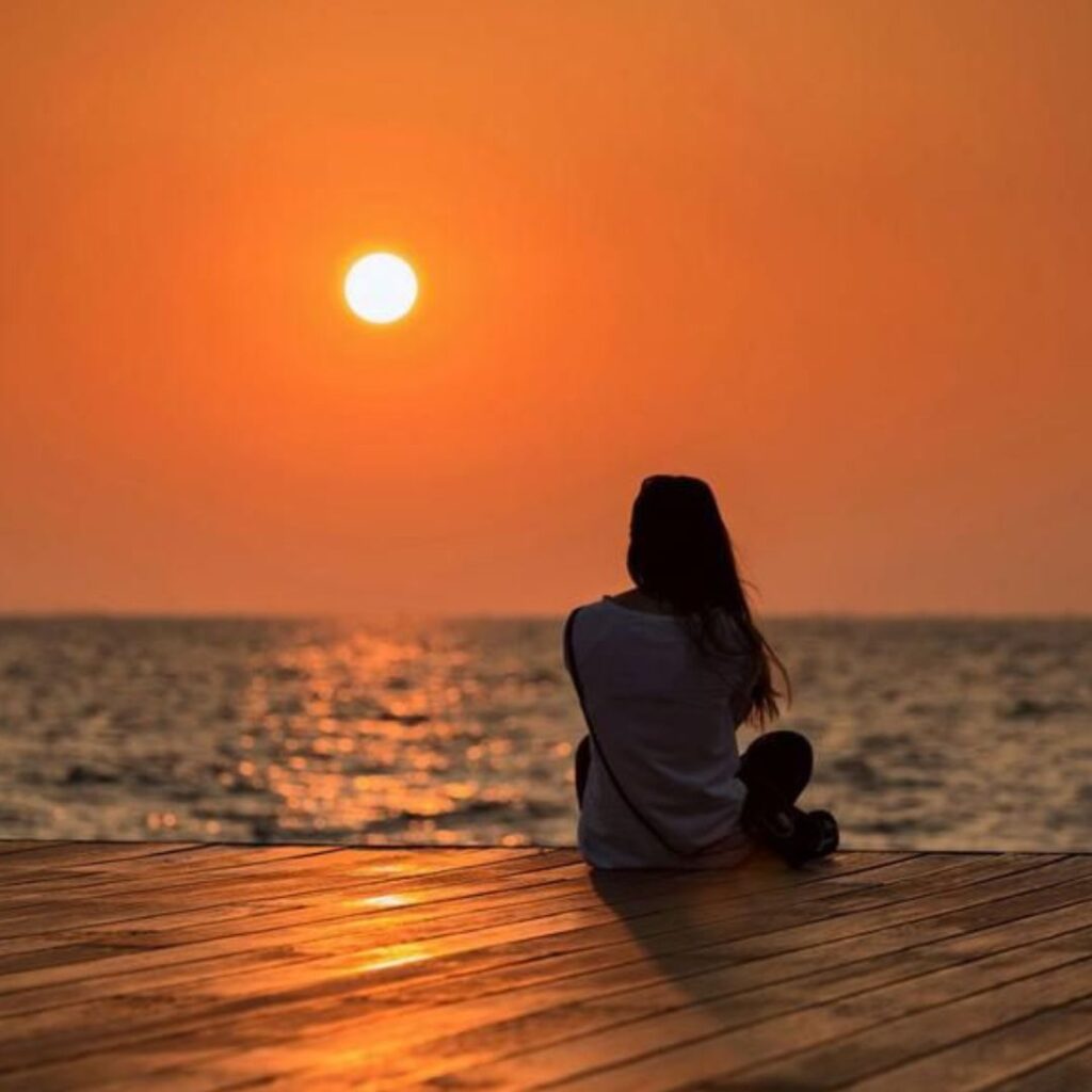 Woman sitting on dock looking into the setting sun and an orange sky